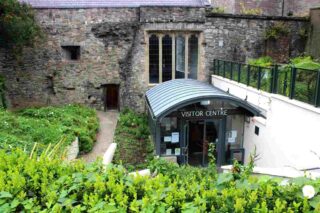 The exterior of the entrance to St Audoen's Visitor Centre. The entry is below street level and the photo is looking down from the main street outside. The doorway is situated in the ancient stone walls of the church. The awning has 'Visitor Centre' written above it. There is lots of greenery to the sides and in front of the entry.