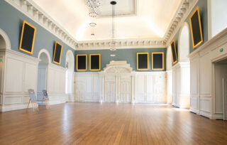 An open room with shiny wooden floorboards, gold frames on the walls, and white designed walls