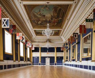 This is a large hall, with crest flags hanging above the mirrors on either side of the room. There are two large chandeliers on the ceiling, and painting on the ceiling in the classical antiquity style